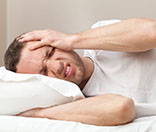 a man wearing a white shirt, laying his head on a white pillow, and holding the left side of his head in chronic pain